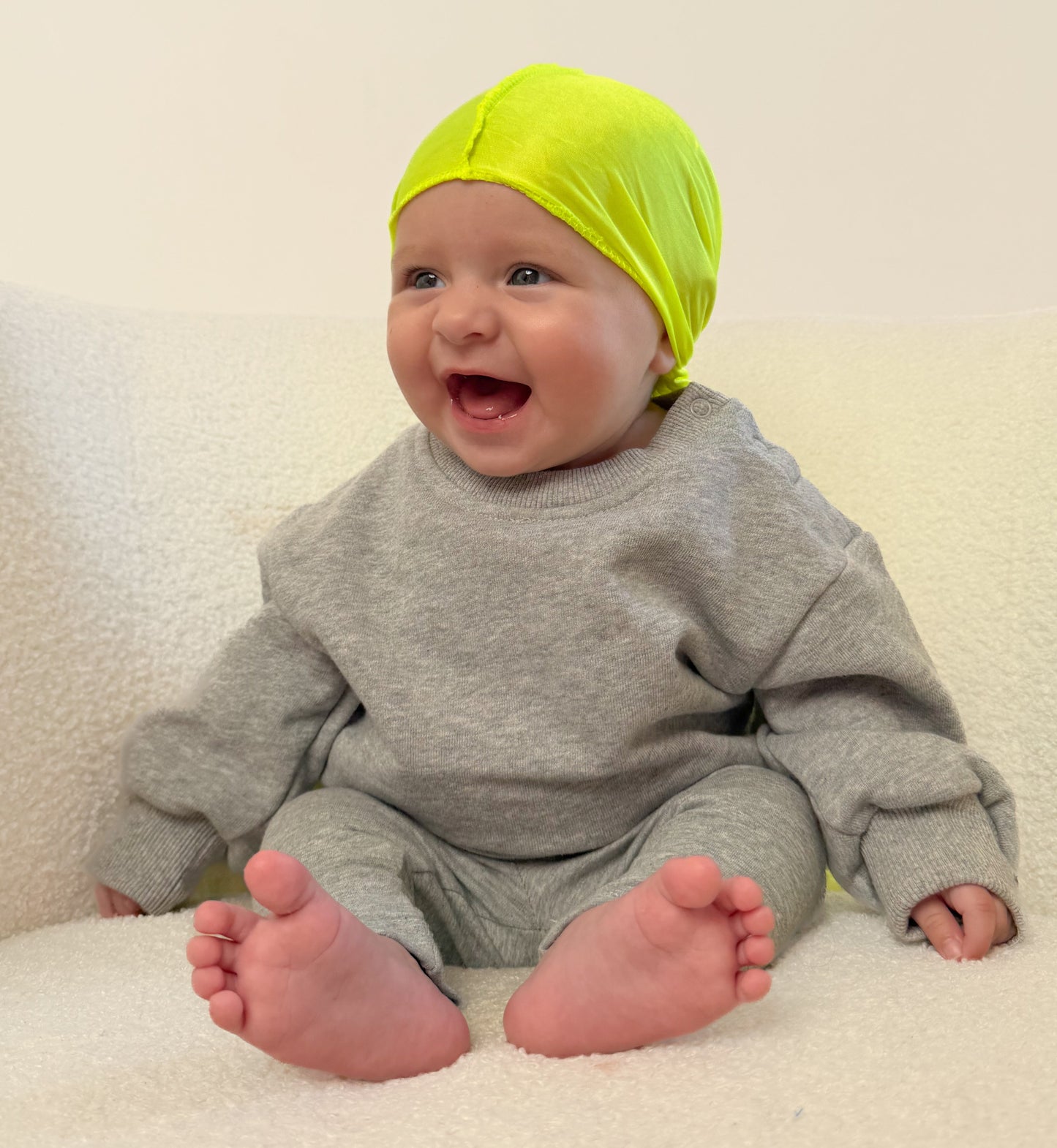 Baby wearing a bright green headband and gray outfit sitting on a white surface.